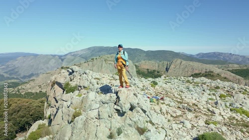Woman with backpack and trekking poles stands on rocky summit overlooking expansive Ponoig (1,181 m.)  mountain summit  landscape and distant Costa Blanca coast - stock video