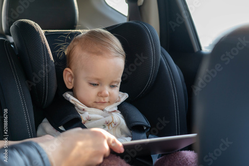фотографія Mother is giving a tablet to her toddler daughter sitting in car seat to enterta