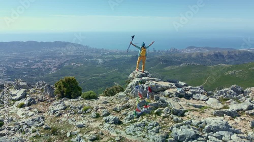 Aerial view of a triumphant hiker standing on the rocky summit of Ponoig (1,181 m.)  mountain with arms raised, celebrating the achievement while overlooking the stunning Costa Blanca coastline.