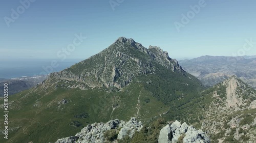 Majestic aerial view moving backward over a rocky ridge, revealing the impressive Puig Campana  (1.406 m) mountain peak against a clear blue sky, with the mediterranean sea visible in the distance.