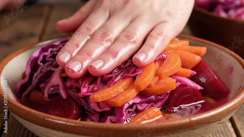 Hand pressing colorful vegetables including red cabbage, carrots, and beets in a wooden bowl on a rustic wooden table, showcasing the preparation of a vibrant salad