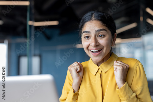 Close-up photo of a young Indian woman sitting at the office desk, looking at the laptop screen and being happy