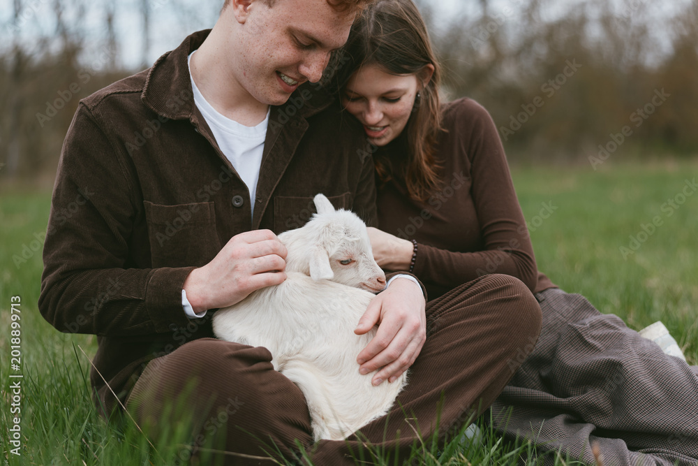 Naklejka premium Couple sits on grass in a meadow with a gentle goat kid resting on their laps, warm affectionate moment outdoors, casual rustic clothing, calm atmosphere, natural light, true lifestyle scene with