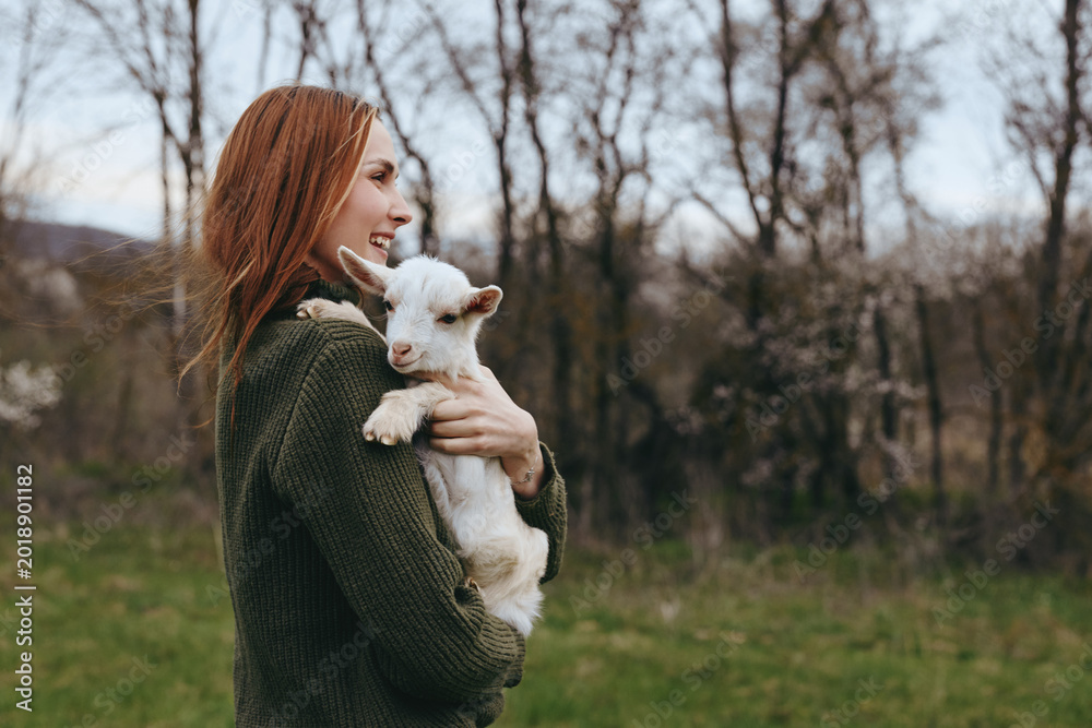 Naklejka premium A woman in a green sweater gently cradles a young goat in a sunlit field, smiling. A candid moment of animal friendship, rural lifestyle, and calm countryside living.