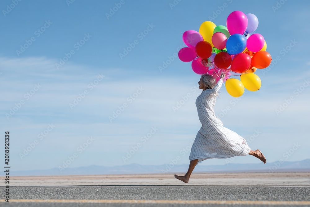 Naklejka premium Energetic woman in flowing white dress leaps barefoot down asphalt road, clutching vibrant balloons against vast desert backdrop under clear blue sky.