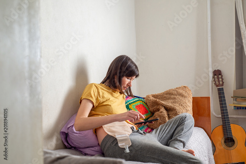 Young woman with a colostomy bag sits on her bed and looks at the phone at home