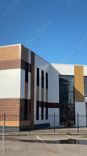 Stylish modern house with siding under blue sky, geometric facade of glass and wood panels, parking lot and metal fence