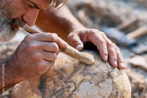 Archaeologist with gray beard carefully cleans ancient pottery artifact using a brush, surrounded by excavation site with soil and tools in the background