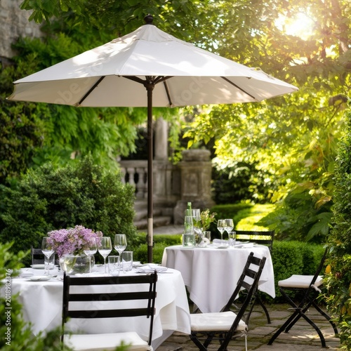 Outdoor Dining Setup With White Tablecloths and an Umbrella Near Green Plants...