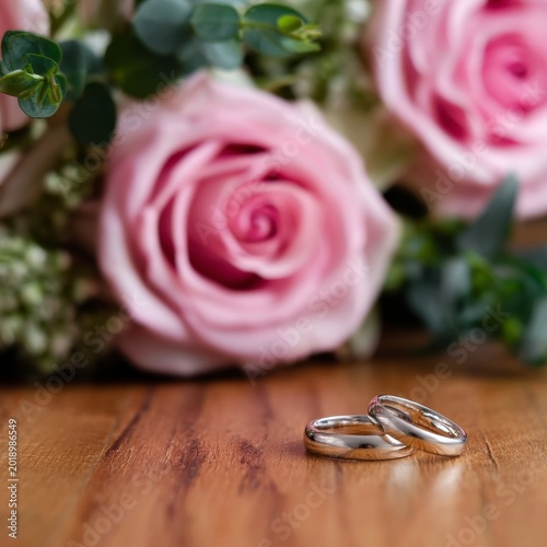 Two Wedding Rings Sit on a Wooden Table Near a Bouquet of Pink Roses With Gre...