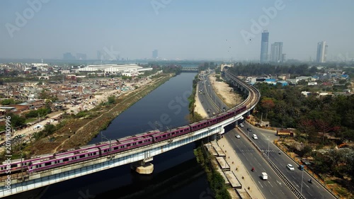 Aerial drone view of two metro trains crossing each other on elevated tracks alongside a river in Delhi city.