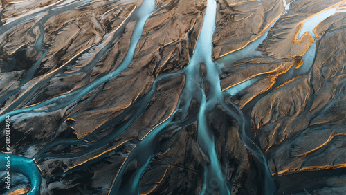 Aerial view of intricate braided glacial river patterns flowing through dark volcanic sand with golden sunlight highlights Skaftarhreppur, Iceland.