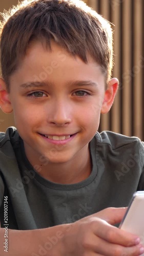 Cheerful boy smiling while holding smartphone outdoors in vertical portrait. Warm sunlight creates a happy childhood lifestyle moment with casual digital interaction in nature. Happy child