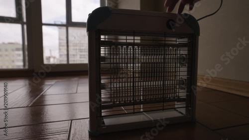 Close-up hand of unrecognizable person places electric bug zapper on wooden floor and turns it on. The ultraviolet lamp glows, ready to attract and kill insects for pest control.