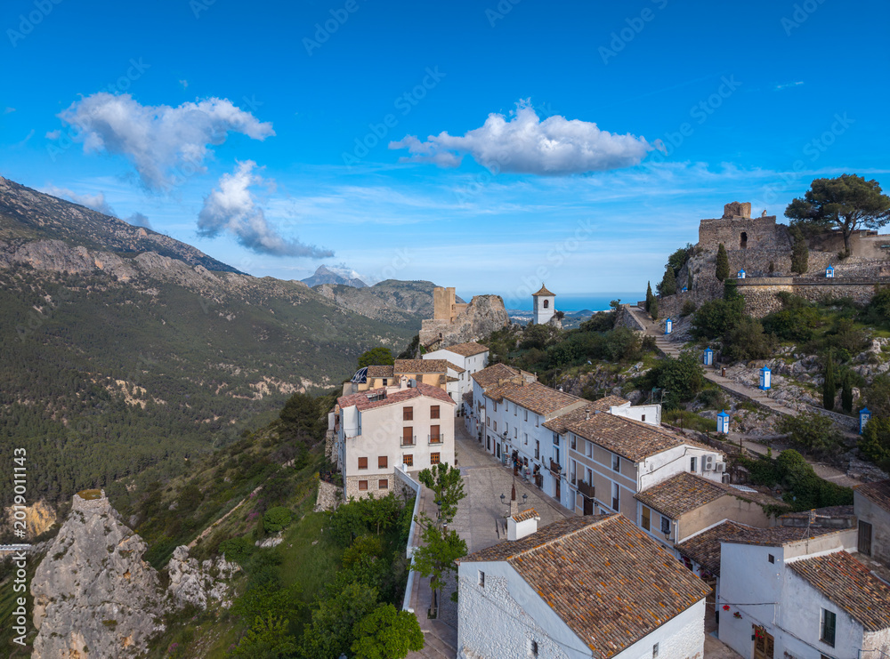 Obraz premium Panoramic view of a picturesque hilltop village in Spain featuring white-washed houses, terracotta rooftops, a medieval castle, and lush green mountains under blue sky