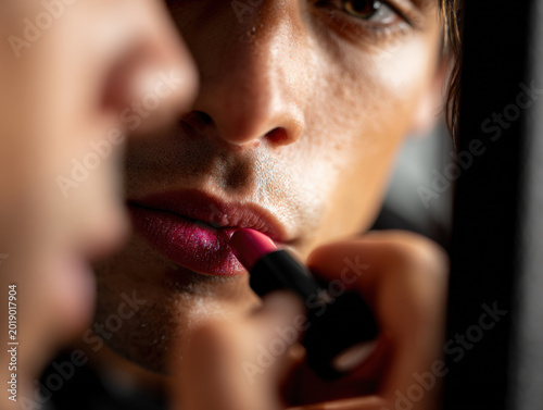 Close-up of man applying lipstick in mirror with focused expression