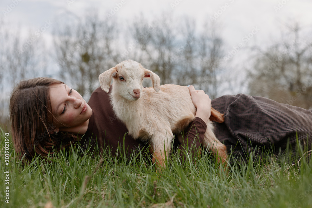 Naklejka premium woman outdoors goat kid lifestyle nature grass smile, cheerful candid moment with a relaxed pose, sunlit scene and natural colors capturing a playful bond in nature