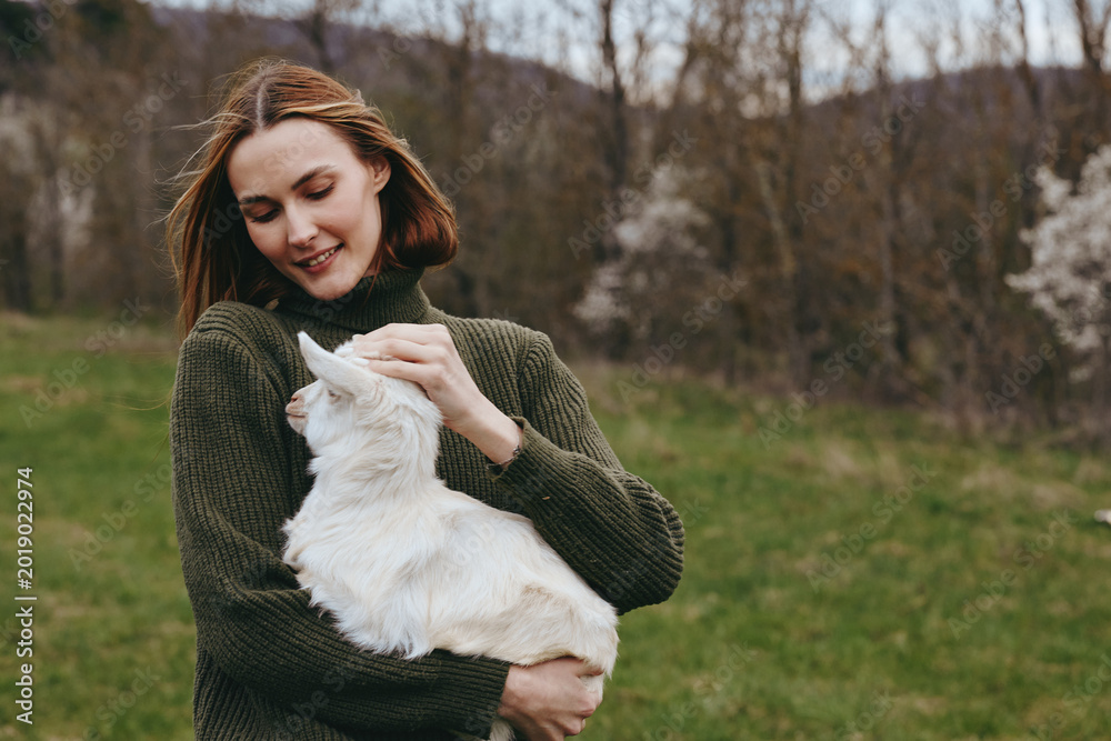 Obraz premium A joyful woman holds a curious goat kid in a sunlit meadow, smiling softly as they share a moment of countryside companionship and rustic lifestyle.