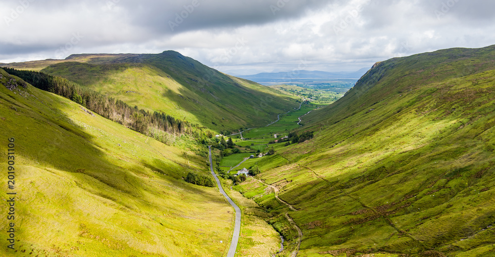 Naklejka premium Aerial view of Glengesh Pass curving through a broad green valley in County Donegal, Ireland. A dramatic birds eye landscape of remote mountains and winding roads