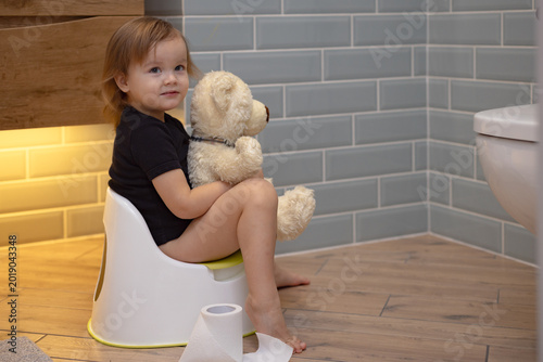 little baby sitting on potty in bathroom. Cute adorable child girl using chamber pot together with her teddy bear. Toddler learning to use the Toilet