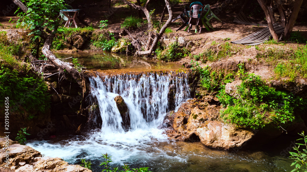Fototapeta premium Small waterfall surrounded by lush vegetation and natural rocks in Akchour Valley, Morocco, with a relaxing riverside resting area.