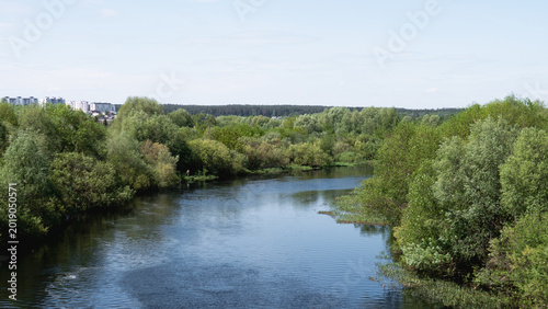 River Flowing Through Lush Green Trees Creating Tranquil Countryside Panorama With Gentle Ripples, Sunlit Sky, Mixed Deciduous Forest Along Meandering Bank, Distant Horizon And Clouds Reflecting