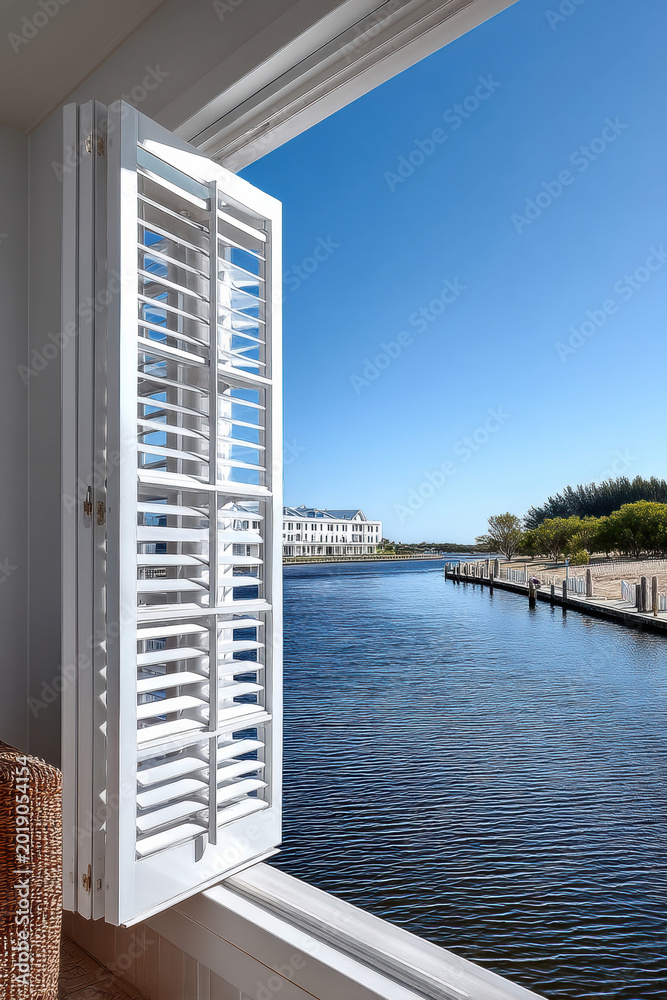 Fototapeta premium White plantation shutters open to tranquil waterfront view, blue sky, calm water, modern architecture, peaceful coastal home