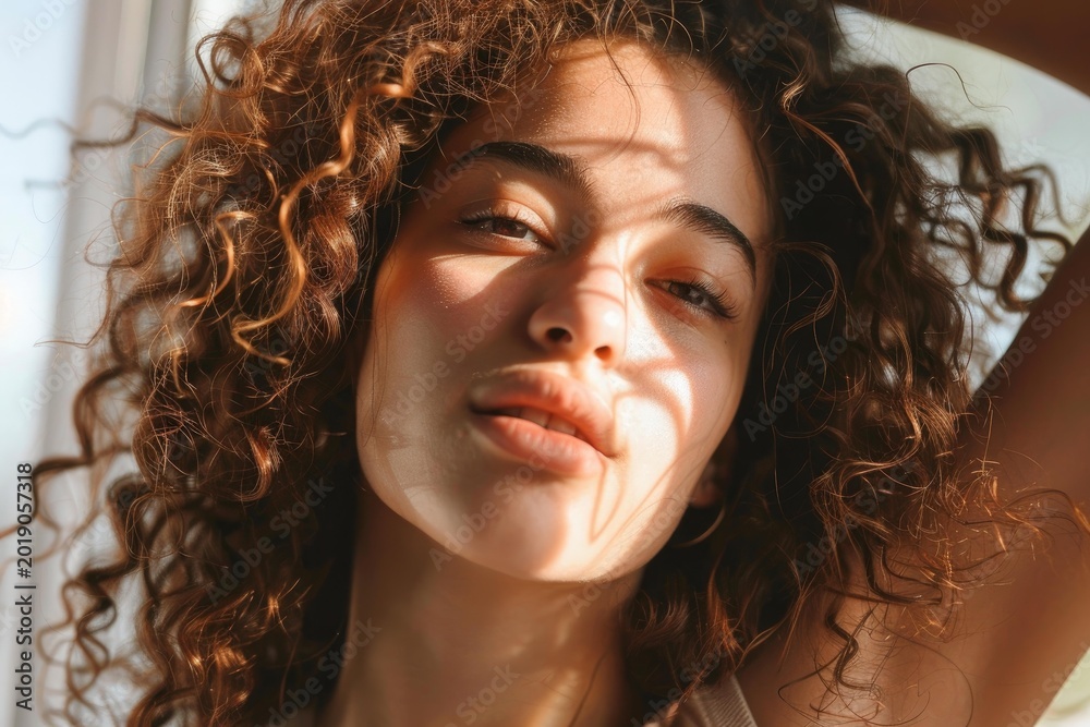 Fototapeta premium Portrait of a beautiful young woman with curly hair posing in sunlight by the window