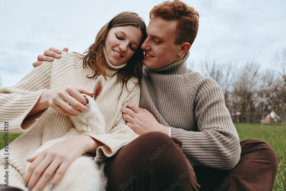 Naklejka premium Smiling couple in chunky sweaters outdoors, caressing a fluffy white dog, capturing a cozy moment of love and companionship. Natural light, soft shadows, rural backdrop, relaxed mood, wellness and