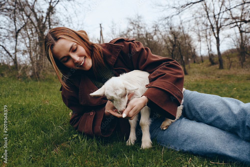 Obraz premium Cheerful woman outdoors, lying on grass with a gentle goat, kid nearby, warm smile, relaxed pose, soft natural light, casual jacket, autumnal park setting, close bond, nature atmosphere