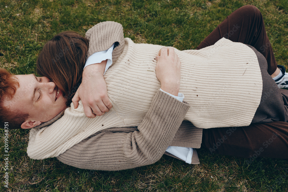 Obraz premium Couple lies on grass, wrapped in cream sweaters, sharing a quiet cuddle in a sunlit park. Cozy, intimate mood emphasizes warmth, affection, and relaxed togetherness.