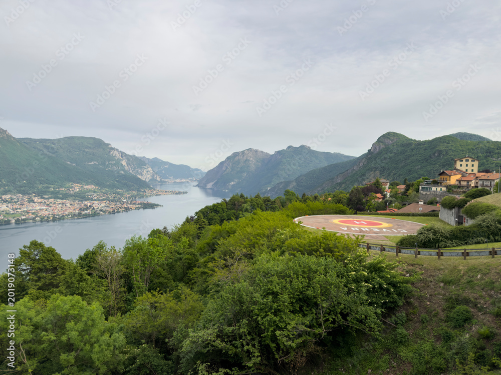 Fototapeta premium Lake Como is stretching through a mountainous landscape, surrounded by lush green trees and a charming village featuring a helipad with civenna written on it