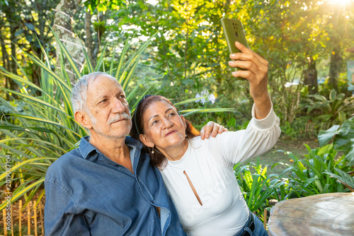 A happy elderly couple take selfies with their cell phone while enjoying time outdoors together in a sunny park