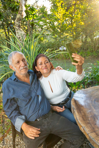 A happy elderly couple take selfies with their cell phone while enjoying time outdoors together in a sunny park