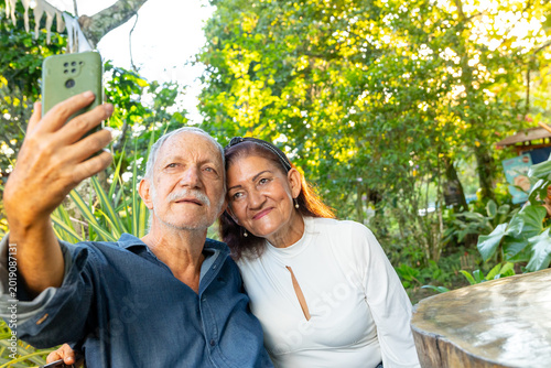 A happy elderly couple take selfies with their cell phone while enjoying time outdoors together in a sunny park