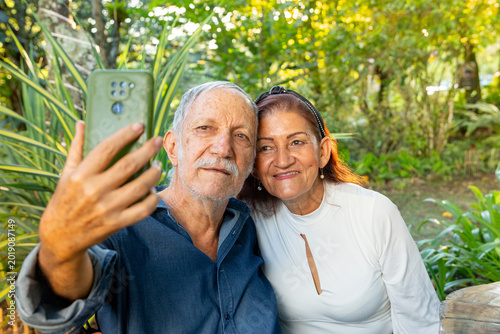 A happy elderly couple take selfies with their cell phone while enjoying time outdoors together in a sunny park