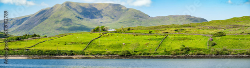 Green mountain slopes rise above Killary Fjord under bright skies in western Ireland. This image is a scenic landscape focused more on the fjord lush hillsides than the water.