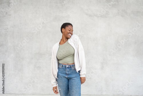 Smiling young woman with eyes closed and raised shoulder, relaxed pose in casual outfit against gray wall with modern everyday lifestyle moment