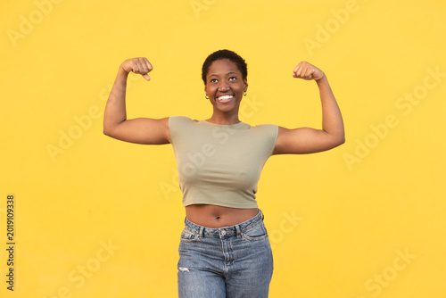 Fit healthy woman flexing muscles against yellow background. Smiling young black lady showing strong body in casual outfit