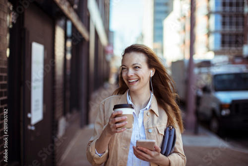 Smiling woman walking in city with coffee and smartphone