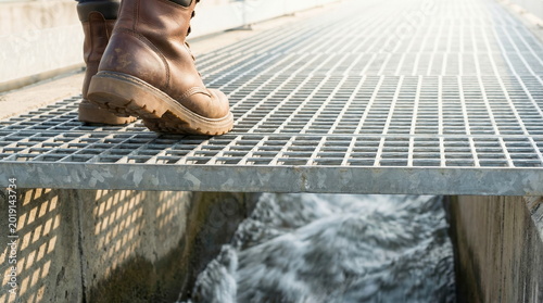 Work boots standing on industrial metal grate above fast-flowing water, symbolizing engineering and infrastructure maintenance.