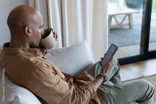 African American man in brown shirt and olive pants sipping mug and scrolling smartphone on sofa