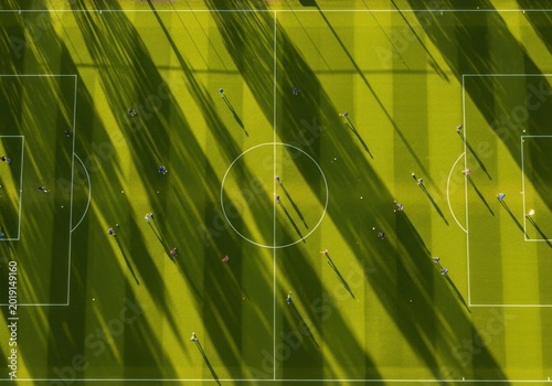 Aerial drone view of professional soccer match on green grass field with long dramatic shadows cast by players during sunny afternoon