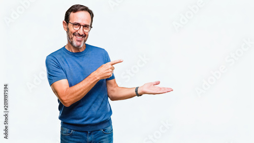 A smiling middle-aged man wearing glasses and casual blue clothing points and presents an empty copy space against a plain white studio background.
