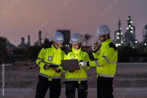 Group of professional petroleum engineers in high-visibility jackets and hardhats working together, using laptop and tablet computers at oil refinery chemical plant industry factory site at twilight.