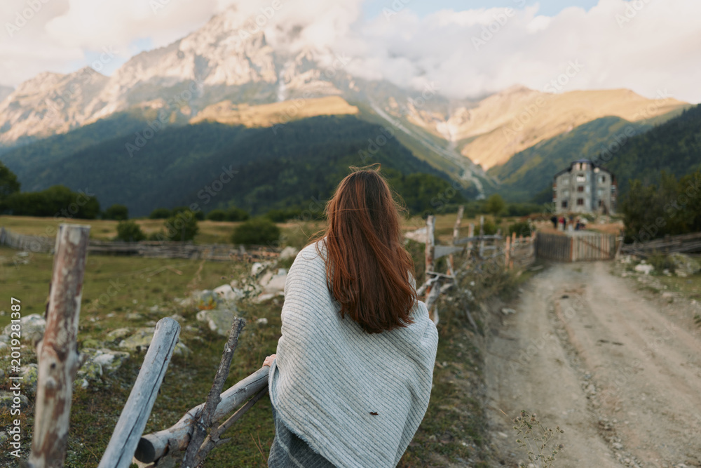 Naklejka premium Young woman with long hair wrapped in blanket stands near wooden fence looks at mountain landscape on sunny day in rural area with dirt road and old house.