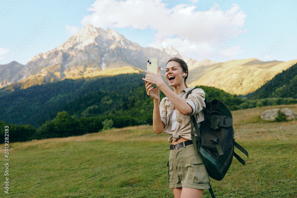 Fototapeta premium Young woman on a hiking trip takes a selfie in a mountain meadow, backpack ready, bright sky, cheerful mood amid vast nature and wildflowers sway nearby under blue sky.
