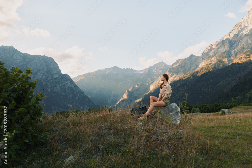 Fototapeta premium Young woman sits on a rock in a meadow with distant mountains, enjoying a peaceful sunny moment.
