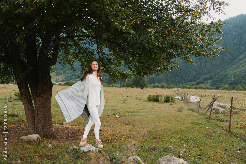 Fototapeta premium A woman in white clothes stands near a tree in a green field with mountains in the background. She holds a light shawl and enjoys the peaceful outdoor lifestyle.