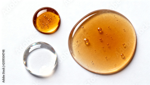 Viscous golden-amber and clear liquid drops arranged in a minimalist abstract composition on a clean white background, highlighting texture, transparency, and fluidity